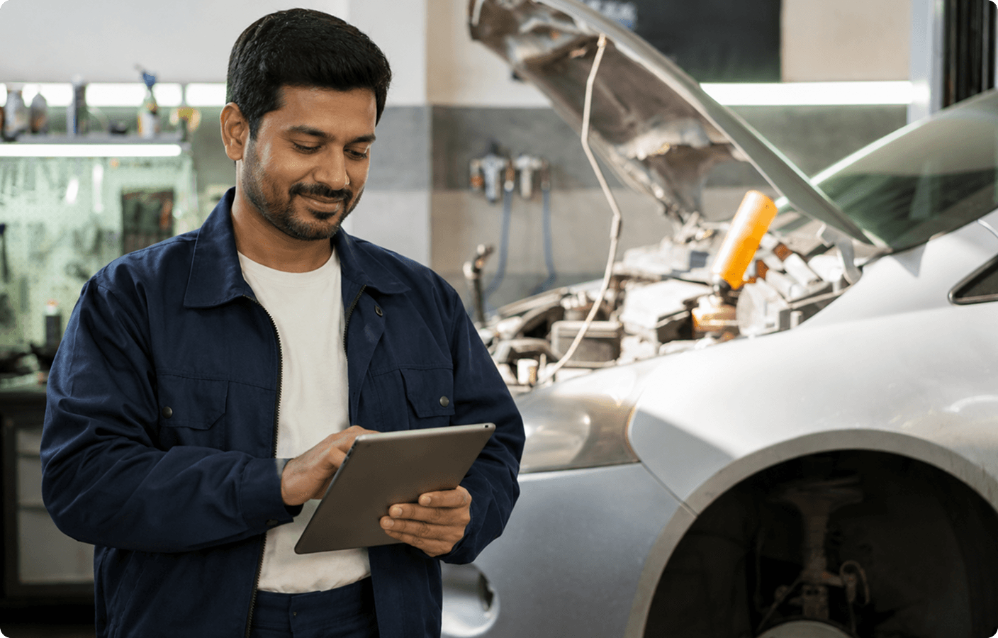 Automobile workshop technician reviewing workforce reports on a tablet beside a car with its hood open