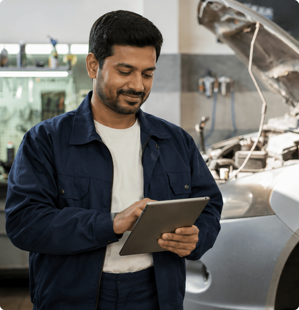 Automobile workshop technician reviewing workforce reports on a tablet beside a car with its hood open