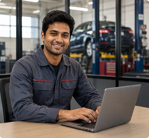 Technician in a blue uniform using a laptop at a desk in an automotive service center