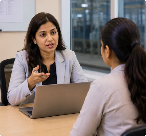 Manager in a light blazer leading a performance review discussion with a colleague at a desk with a laptop