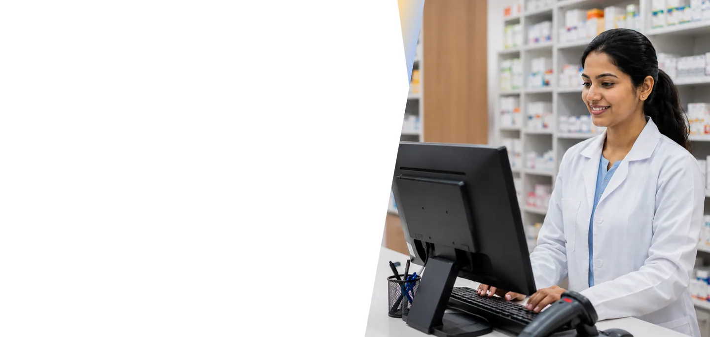 Pharmacist in a white lab coat working at a computer in a pharmacy with medication shelves in the background